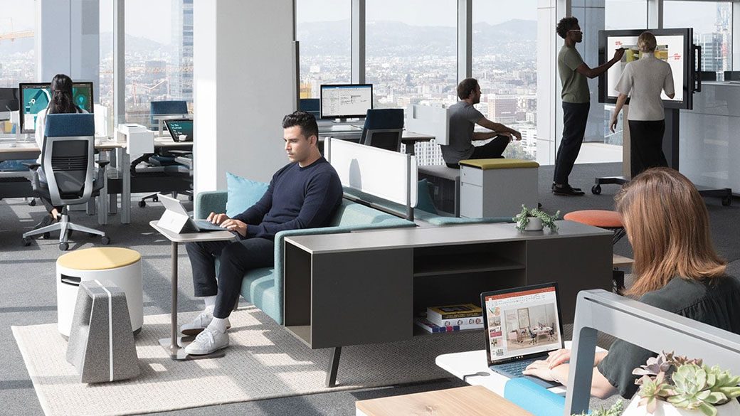 A modern workspace, featuring coworkers on various Surface devices: a man types on a Surface Pro, a woman types on Surface Laptop, and a man and a woman use a Surface Hub.